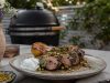 Sliced herb-crusted grilled beef on a plate with a lemon half, white sauce dollop, and pistachio crumble, outdoors near a grill.
