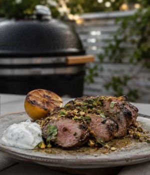 Grillad-lammytterfile2 Sliced herb-crusted grilled beef on a plate with a lemon half, white sauce dollop, and pistachio crumble, outdoors near a grill.