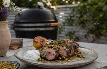 Sliced herb-crusted grilled beef on a plate with a lemon half, white sauce dollop, and pistachio crumble, outdoors near a grill.