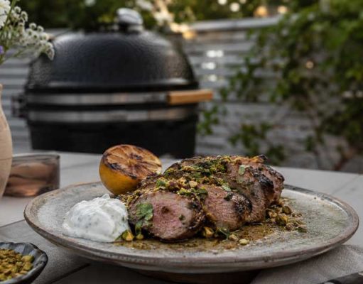 Grillad lammytterfilé med pistage, mynta och grillad citron Sliced herb-crusted grilled beef on a plate with a lemon half, white sauce dollop, and pistachio crumble, outdoors near a grill.