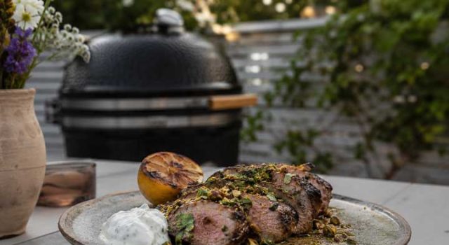 Sliced herb-crusted grilled beef on a plate with a lemon half, white sauce dollop, and pistachio crumble, outdoors near a grill.