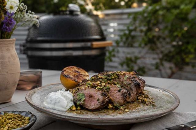 Sliced herb-crusted grilled beef on a plate with a lemon half, white sauce dollop, and pistachio crumble, outdoors near a grill.
