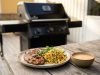 Grilled chicken with corn salsa, lime wedges, and cilantro on a plate beside a bowl of sauce on a wooden table outdoors; barbecue grill in background.
