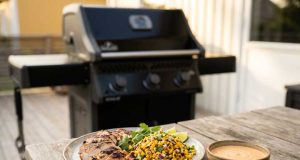 Kycklinglårfilé på grillen med rostad majs-salsa och chipotle-lime Grilled chicken with corn salsa, lime wedges, and cilantro on a plate beside a bowl of sauce on a wooden table outdoors; barbecue grill in background.