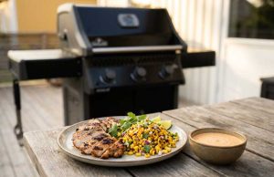 Kycklinglårfilé på grillen med rostad majs-salsa och chipotle-lime Grilled chicken with corn salsa, lime wedges, and cilantro on a plate beside a bowl of sauce on a wooden table outdoors; barbecue grill in background.