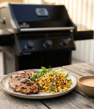 KYCKLI~2 Grilled chicken with corn salsa, lime wedges, and cilantro on a plate beside a bowl of sauce on a wooden table outdoors; barbecue grill in background.