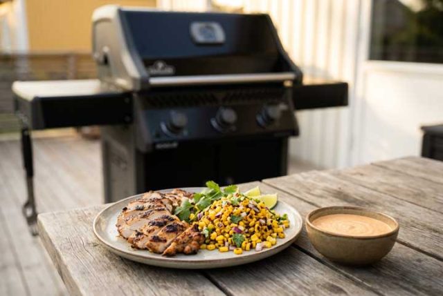 Grilled chicken with corn salsa, lime wedges, and cilantro on a plate beside a bowl of sauce on a wooden table outdoors; barbecue grill in background.