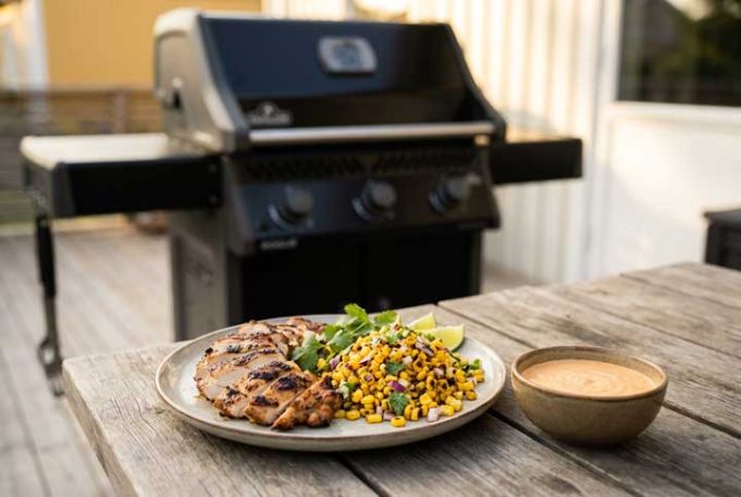 Grilled chicken with corn salsa, lime wedges, and cilantro on a plate beside a bowl of sauce on a wooden table outdoors; barbecue grill in background.