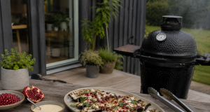 Platter of grilled eggplant slices topped with cheese, herbs, and pomegranate seeds on a wooden outdoor table beside a smoker.