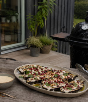 Platter of grilled eggplant slices topped with cheese, herbs, and pomegranate seeds on a wooden outdoor table beside a smoker.