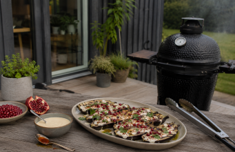Rökbakad aubergine med tahini, granatäpple och chiliolja Platter of grilled eggplant slices topped with cheese, herbs, and pomegranate seeds on a wooden outdoor table beside a smoker.