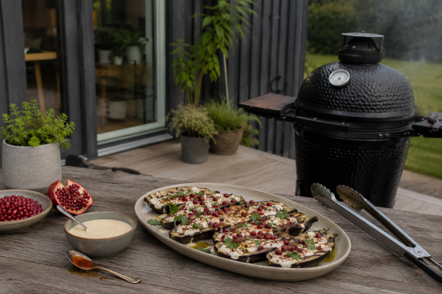 Platter of grilled eggplant slices topped with cheese, herbs, and pomegranate seeds on a wooden outdoor table beside a smoker.
