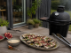 Rökbakad aubergine med tahini, granatäpple och chiliolja Platter of grilled eggplant slices topped with cheese, herbs, and pomegranate seeds on a wooden outdoor table beside a smoker.