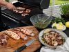 Outdoor barbecue scene with a cutting board of grilled seafood and meat, a knife, and bowls of salsa, lemon wedges, and herbs on a patio table.