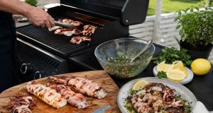 Outdoor barbecue scene with a cutting board of grilled seafood and meat, a knife, and bowls of salsa, lemon wedges, and herbs on a patio table.