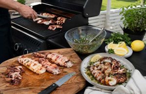 Outdoor barbecue scene with a cutting board of grilled seafood and meat, a knife, and bowls of salsa, lemon wedges, and herbs on a patio table.