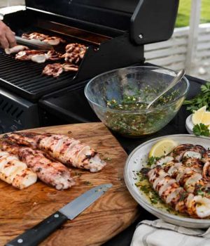 Outdoor barbecue scene with a cutting board of grilled seafood and meat, a knife, and bowls of salsa, lemon wedges, and herbs on a patio table.