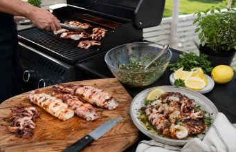 Outdoor barbecue scene with a cutting board of grilled seafood and meat, a knife, and bowls of salsa, lemon wedges, and herbs on a patio table.