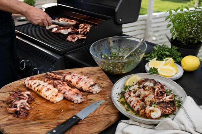 Grillad bläckfisk med citron, vitlök och persilja Outdoor barbecue scene with a cutting board of grilled seafood and meat, a knife, and bowls of salsa, lemon wedges, and herbs on a patio table.