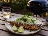 Grilled whole fish with sesame and herbs on a plate, lime wedges and cucumber salad on the side.