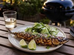 Grilled whole fish with sesame and herbs on a plate, lime wedges and cucumber salad on the side.