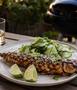Grilled whole fish with sesame and herbs on a plate, lime wedges and cucumber salad on the side.