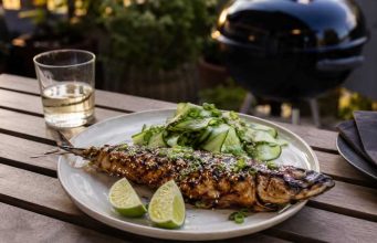 Grilled whole fish with sesame and herbs on a plate, lime wedges and cucumber salad on the side.