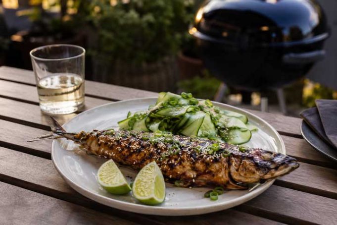 Grilled whole fish with sesame and herbs on a plate, lime wedges and cucumber salad on the side.