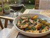 Grillad Potatissallad med Kapris, Dill och Senapsvinägrett Bowl of roasted potato wedges with dill on a wooden outdoor table, barbecue in the background
