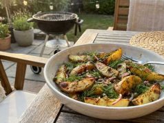 Grillad Potatissallad med Kapris, Dill och Senapsvinägrett Bowl of roasted potato wedges with dill on a wooden outdoor table, barbecue in the background