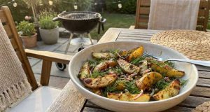Grillad Potatissallad med Kapris, Dill och Senapsvinägrett Bowl of roasted potato wedges with dill on a wooden outdoor table, barbecue in the background