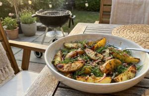 Grillad Potatissallad med Kapris, Dill och Senapsvinägrett Bowl of roasted potato wedges with dill on a wooden outdoor table, barbecue in the background