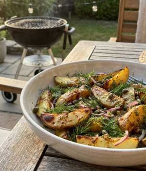 Bowl of roasted potato wedges with dill on a wooden outdoor table, barbecue in the background
