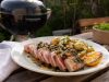 Grillad tonfisk med kapris, brynt smör och sotad citron Plate of sliced seared tuna steak with pink center, roasted potatoes, lemon wedge, and herb garnish on a wooden table outdoors. A grill is in the background.