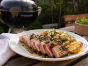 Grillad tonfisk med kapris, brynt smör och sotad citron Plate of sliced seared tuna steak with pink center, roasted potatoes, lemon wedge, and herb garnish on a wooden table outdoors. A grill is in the background.