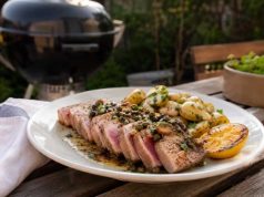 Plate of sliced seared tuna steak with pink center, roasted potatoes, lemon wedge, and herb garnish on a wooden table outdoors. A grill is in the background.