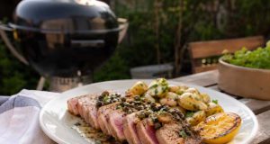 Grillad tonfisk med kapris, brynt smör och sotad citron Plate of sliced seared tuna steak with pink center, roasted potatoes, lemon wedge, and herb garnish on a wooden table outdoors. A grill is in the background.