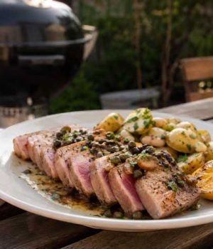 Plate of sliced seared tuna steak with pink center, roasted potatoes, lemon wedge, and herb garnish on a wooden table outdoors. A grill is in the background.
