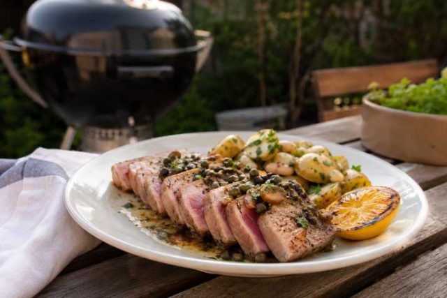 Plate of sliced seared tuna steak with pink center, roasted potatoes, lemon wedge, and herb garnish on a wooden table outdoors. A grill is in the background.