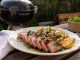 Plate of sliced seared tuna steak with pink center, roasted potatoes, lemon wedge, and herb garnish on a wooden table outdoors. A grill is in the background.