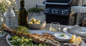 Whole grilled fish on a platter with cucumber salad, lemon wedges, and herb yogurt sauce, on an outdoor table with a grill in the background.