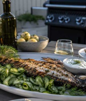 Whole grilled fish on a platter with cucumber salad, lemon wedges, and herb yogurt sauce, on an outdoor table with a grill in the background.