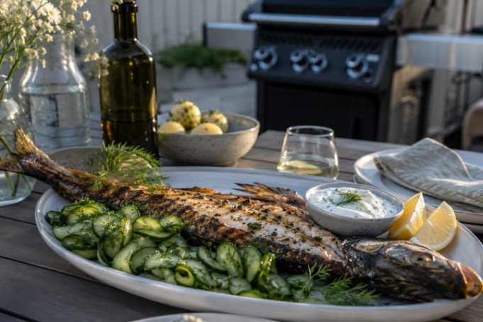 Whole grilled fish on a platter with cucumber salad, lemon wedges, and herb yogurt sauce, on an outdoor table with a grill in the background.