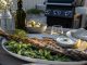 Whole grilled fish on a platter with cucumber salad, lemon wedges, and herb yogurt sauce, on an outdoor table with a grill in the background.