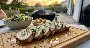 Fläskytterfile med Vitlökssmör och Örter Herb-garnished sliced pork tenderloin on a wooden board, with a side salad and roasted potatoes, outdoor sunset patio.