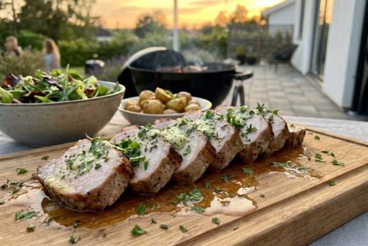 Herb-garnished sliced pork tenderloin on a wooden board, with a side salad and roasted potatoes, outdoor sunset patio.