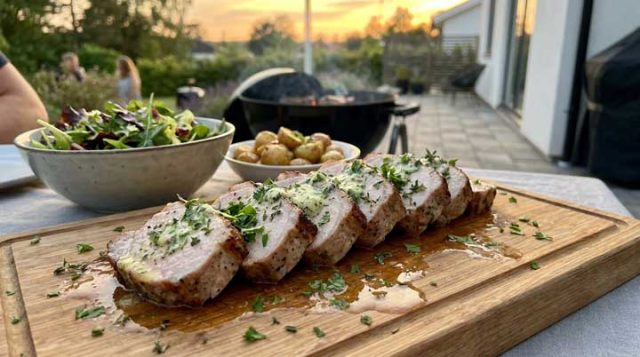 Herb-garnished sliced pork tenderloin on a wooden board, with a side salad and roasted potatoes, outdoor sunset patio.