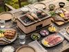 Outdoor picnic-style grill master on a wooden table with skewers cooking; surrounding plates hold meat, fish, vegetables, lemon, and condiments.