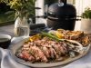 Sliced medium-rare steak on a platter with lemon wedges, grilled bread, and asparagus, outdoors near a barbecue grill.