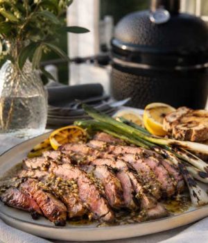pluma-grillen-04-fardig-ratt Sliced medium-rare steak on a platter with lemon wedges, grilled bread, and asparagus, outdoors near a barbecue grill.